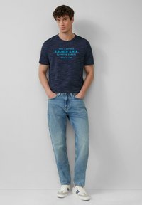 Donkerblauw heather t-shirt met contrasterende turquoise tekst, gecombineerd met lichtblauwe jeans en witte sneakers. Casual pasvorm en design.