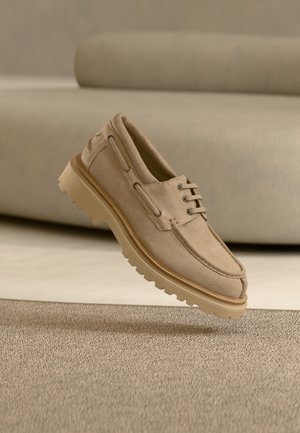 Beige mokkanahkainen rento nauhallinen kenkä, jossa paksu uurrettu pohja, esitetty ilmassa sen kanssa yhteensopivan beigen tekstuurisen pinnan ja taustan yläpuolella.