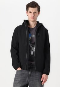 Casaco softshell preto com fecho éclair na frente, dois bolsos laterais e capuz. Usado sobre uma t-shirt gráfica escura e jeans cinzentos. Textura suave.