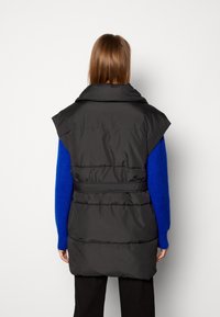 Person med långt brunt hår som bär en svart vadderad ärmlös väst över en blå tröja och svarta byxor, sedd bakifrån mot en vit bakgrund.