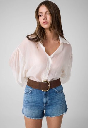 Junge Frau mit langen braunen Haaren trägt eine lockere weiße Bluse mit Knöpfen, einen braunen Gürtel und blaue Jeansshorts vor einem einfarbigen Hintergrund.
