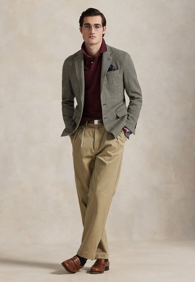 Homme portant des lunettes, blazer gris, polo bordeaux, pantalon beige, chaussures en cuir marron, debout les mains dans les poches devant un fond uni.