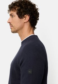 Navy blue knit sweater with a round neck, textured fabric, and ribbed cuffs. Features a small black logo patch on the left sleeve.