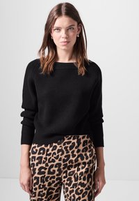 Schwarzer, gerippter Pullover mit weitem Ausschnitt und langen Ärmeln, kombiniert mit beigen Hosen, die mit einem auffälligen schwarzen Leopardenmuster versehen sind.