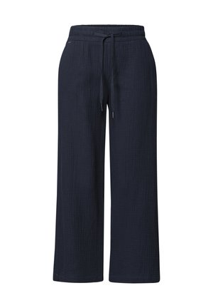 Femme portant une veste en jean claire et un pantalon bleu marine à cordon, debout contre un mur blanc texturé.