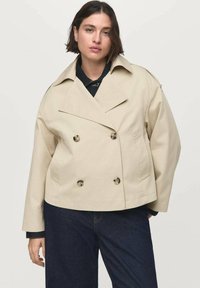 Beige dubbelknäppt jacka med en bred krage, knappar fram och två sidofickor. Buren över en mörk outfit.