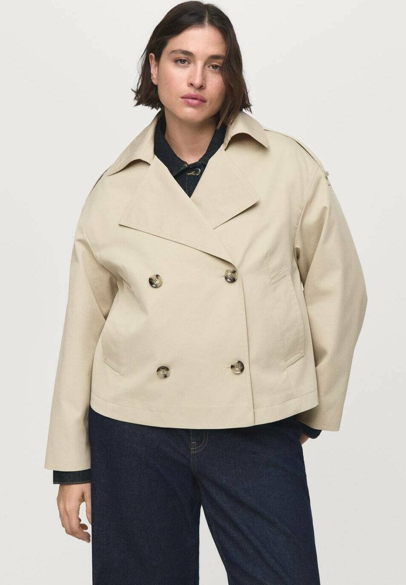 Beige dubbelknäppt jacka med en bred krage, knappar fram och två sidofickor. Buren över en mörk outfit.