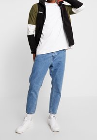 Giacca nera e verde oliva con zip e maniche a colori a blocchi, t-shirt bianca, jeans azzurri chiari e sneakers bianche con suola testurizzata.
