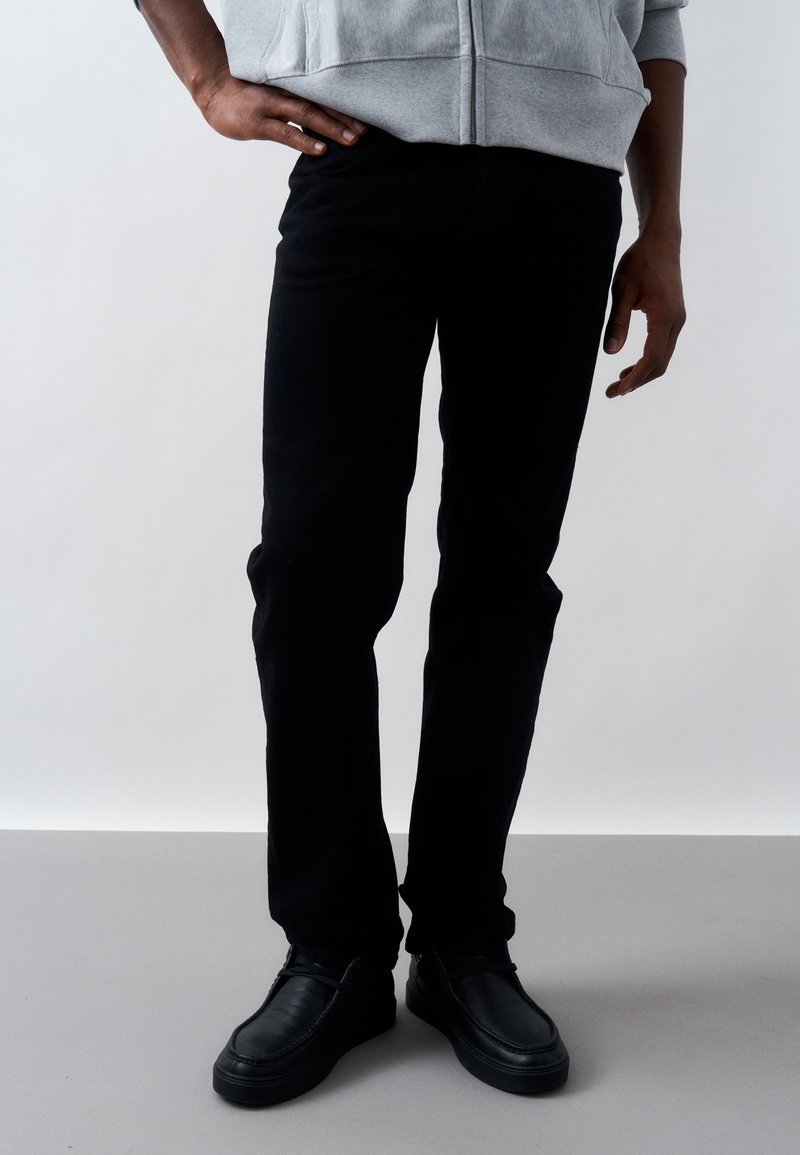 Άτομο που στέκεται φορώντας μαύρο παντελόνι, μαύρα μοκασίνια και ανοιχτό γκρι φούτερ με τα χέρια χαλαρά στο πλάι, πάνω σε απλό φόντο.