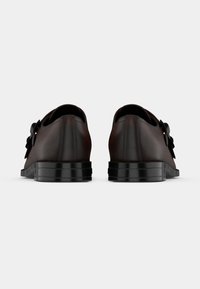 Pāris tumši brūnu ādas oficiālo apavu ar melnām zolēm, skatīti no aizmugures uz vienkārša gaiša fona.