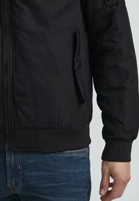 Crna bomber jakna s prednjim zatvaračem, rebrastim manžetama i rubom, te bočnim džepovima. Materijal je gladak s mat završetkom.