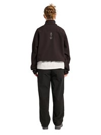 Personne aux cheveux tressés porte une veste noire avec le texte "ATW" dans le dos, une chemise blanche, un pantalon noir et des chaussures grises, debout, tournée dans le dos.