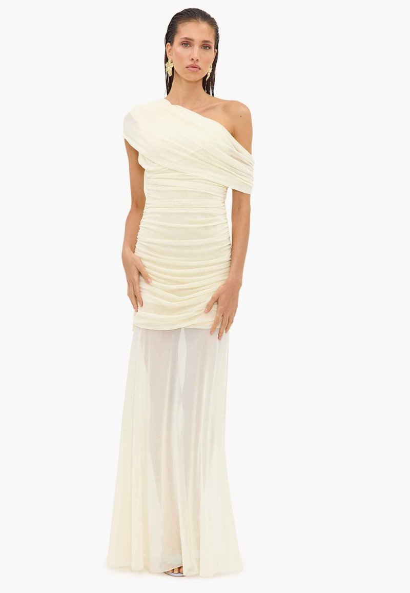 Femme portant une robe crème asymétrique froncée avec une jupe longue transparente et des boucles d'oreilles dorées imposantes, debout devant un fond uni.
