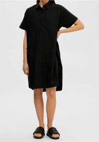 Svart skjortklänning med korta ärmar, avslappnad passform och krage. Tyget verkar lätt och ventilerat, kombinerat med svarta slide-sandaler.