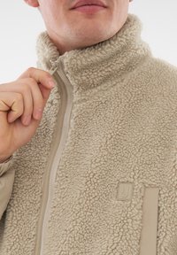 Beige fleece jakke med høj krave, struktureret stof og lynlås foran. Har et logo patch på brystet og kontrasterende kant.