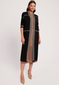 Czarny długi kardigan z białymi detalami przeszycia, zestawiony z dopasowaną brązową sukienką. Tkanina ma gładką fakturę, a całość uzupełniają beżowe szpilki w szpic.