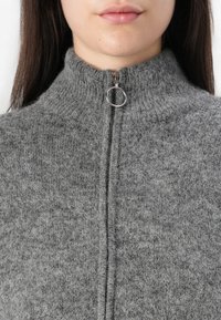 Szary sweter dzianinowy z wysokim kołnierzem, posiadający srebrny zamek błyskawiczny z okrągłym uchwytem. Miękka faktura, średnia waga i dopasowany krój.