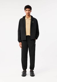 Juoda zip-up striukė su gobtuvu, kreminės spalvos polo marškiniai po ja, juodos sportinės kelnės ir tamsūs sportiniai bateliai su tekstūruotais padais.