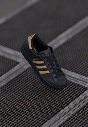 Baskets Adidas Superstar noires avec des bandes beiges et une languette au talon, positionnées au-dessus d'un sol en grille métallique.