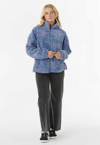 Niebieska kurtka polarowa z zamkiem z przodu, zdobiona teksturowanym wzorem liści. Do tego ciemnoszare spodnie i czarne trampki.