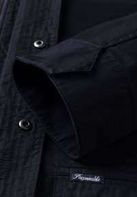 Camisa negra de manga larga con puño doblado, botones de presión metálicos visibles y etiqueta "Façonnable" en el área del bolsillo.