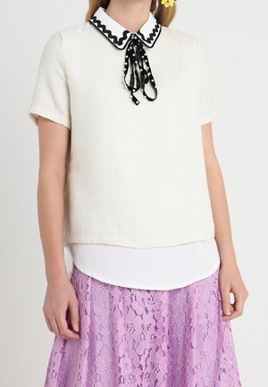 Vrouw draagt een wit shirt met korte mouwen, een zwart-witte polkadotstrik en een lila kanten rok.
