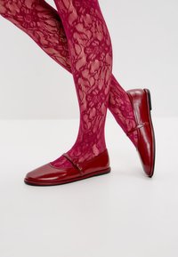 Bailarinas de charol rojo con puntera redonda, que cuentan con una tira decorativa y un hebilla. Llevadas con medias de encaje fucsias con estampado.