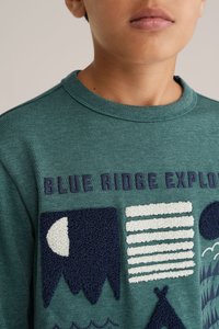 Zeleni pulover z teksturo, ki vključuje napis "BLUE RIDGE EXPLORE" ter različne temno modre in bele vzorce, vključno z gorami, črtami in oblikami.