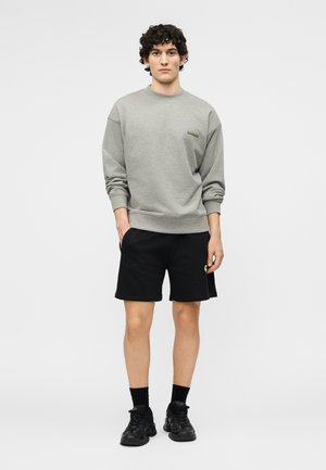 Joven con cabello rizado, vestido con sudadera gris, pantalones cortos negros, calcetines negros y zapatillas negras, posando frente a un fondo blanco liso.