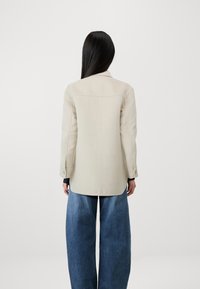 Chaqueta entallada color beige con un diseño estructurado; mangas largas, puños de un solo botón y espalda dividida. Combinada con pantalones vaqueros de pierna ancha en azul.