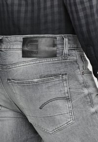 Los jeans de mezclilla gris cuentan con un parche de cuero con el logo en la cintura, un diseño de cinco bolsillos y un sutil desgaste. Se llevan con una camisa a cuadros.