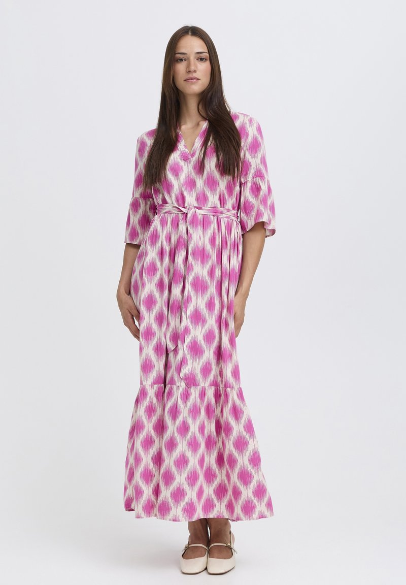 Femme aux longs cheveux bruns portant une robe maxi ceinturée à motifs roses et blancs et des chaussures plates crème, debout devant un fond uni.