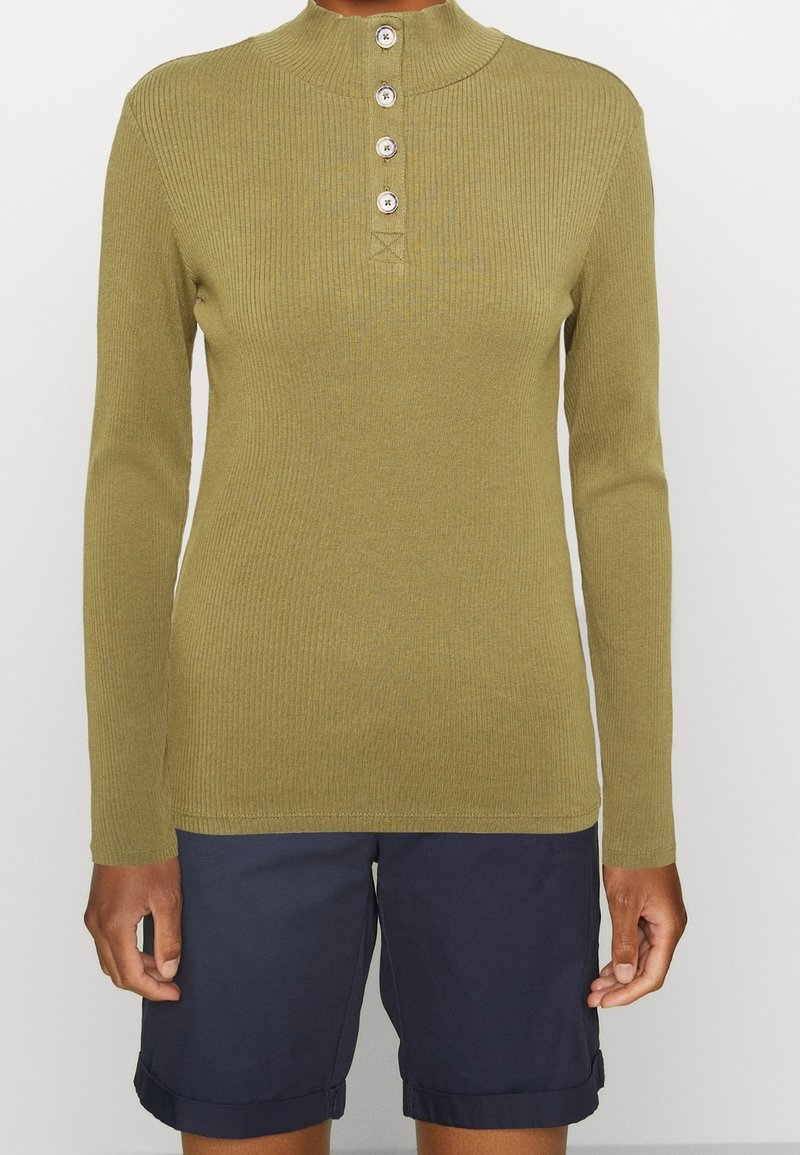 Maglia lungo a maniche con coste di colore verde oliva, colletto alto e pattina con quattro bottoni. Tessuto morbido, design aderente, abbinata a pantaloni corti blu navy.