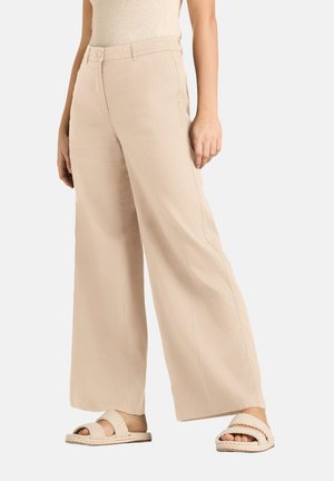 Person, die beige weite Hosen und passende gewebte Slidesandalen trägt, steht vor einem schlichten hellen Hintergrund.