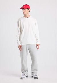 Weißes langärmliges Hemd, graue Jogginghose, roter Hut. Lässiges Outfit mit weichen Texturen und entspannter Passform. Sneakers mit grauen und blauen Akzenten.