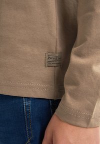 Camicetta beige a maniche lunghe con scollo rotondo, realizzata in cotone, con una piccola etichetta con la scritta "Petrol Ind" e dettagli di cucitura visibili.
