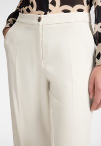 Pantalones tailored de color crema, con una textura suave, que cuentan con un cierre de botón y bolsillos laterales, mostrados con una blusa de estampado.