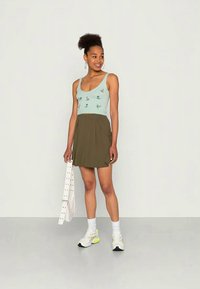 Top azul claro sem mangas com bordado floral, combinado com uma saia plissada verde escura. A modelo usa ténis brancos e segura uma bolsa branca com tachas.