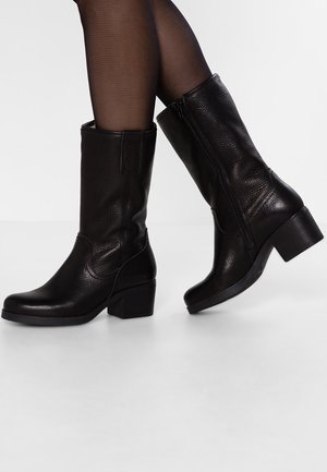 Czarne skórzane botki sięgające połowy łydki na słupkowym obcasie, noszone z przezroczystymi czarnymi rajstopami, prawa stopa lekko uniesiona, pokazująca boczny zamek.