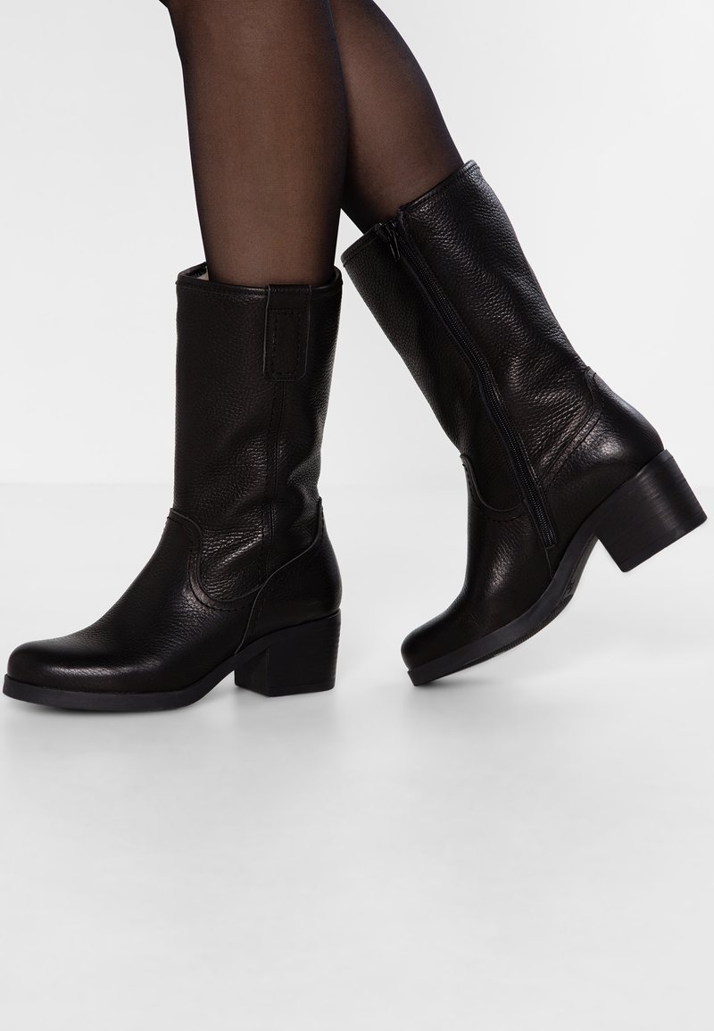 Bottes noires en cuir mi-mollet à talons carrés portées avec des collants noirs transparents, pied droit légèrement levé montrant la fermeture éclair latérale.