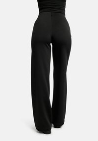 Osoba oblečená v čiernych nohaviciach s vysokým pásom a širokými nohavicami a čiernych topánkach, zobrazená zospodu na jednoduchom svetlom pozadí.