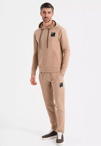 Beige bomullshuvtröja med dragsko, framficka, kombinerad med matchande joggers som har sidfickor och logodetaljer. Bruna sneakers.