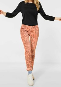 Smala byxor i en texturerad korallfärg med ett bladmönster, kombinerat med en svart långärmad topp och vita sneakers.