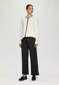 Witte blouse met zwarte bies, lange mouwen en een keyhole halslijn, gecombineerd met brede zwarte broeken en zwarte gepolijste schoenen.