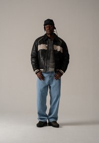 Black leather bomber jacket with embroidered lettering, gray shirt beneath, light blue straight-leg jeans, black shoes, and a black bucket hat.
