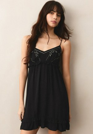 Jeune femme aux longs cheveux foncés portant une robe noire sans manches avec de fines bretelles et une broderie florale subtile.