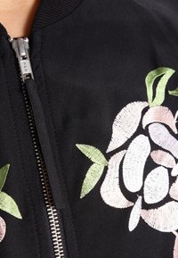 Svart bomberjacka med blommig broderi i rosa och grönt. Dragkedja framtill med en silverfärgad metalldragkedja. Slät yta.