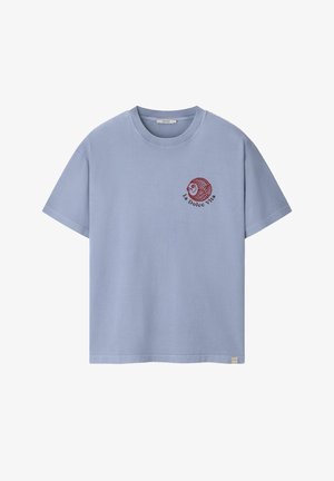 Man met krullend haar die een lichtblauw oversized T-shirt met korte mouwen draagt, gezien van achteren tegen een beige achtergrond.