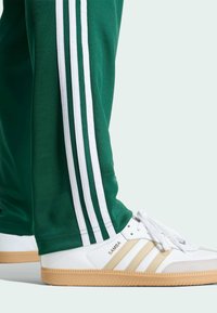 Zelené tepláky s bílým designem tří pruhů, doplněné o bílé tenisky s zlatými akcenty a texturovanou gumovou podrážkou, označené "SAMBA".