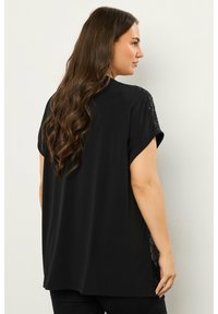 Weit geschnittenes schwarzes Oberteil mit umgeschlagenen Ärmeln, verziert mit strukturierten schwarzen Spitzenakzenten an den Schultern und seitlichen Schlitzen, aus weichem Stoff gefertigt.